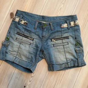 Unika jeansshorts med detaljer - Snygga blå jeansshorts med coola gröna knappar, dragkedjor framtill. Har ett mönstrat tyg vid linningen och tryckt text baktill.
