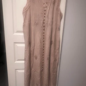 Beige långklänning med broderi och knappar - Långklänning i beige med broderade detaljer och dekorativa knappar framtill. Ärmlös modell med slits nertill och knytband i ryggen. Tunt, mjukt tyg som ger en avslappnad och bohemisk känsla.100%viskos 