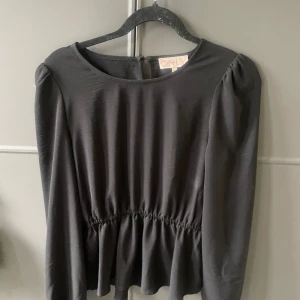 Svart blus med markerad midja från ONLY - Supersnygg svart blus från ONLY med rund halsringning, puffiga axlar och lång ärm. Midjan är markerad med resår för en feminin siluett och blusen har lätt volang nertill. Perfekt att styla med jeans eller skinnbyxor för en trendig look.