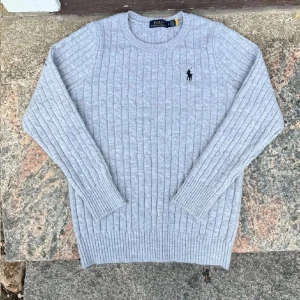 Grå kabelstickad tröja Polo Ralph Lauren - Kabelstickad grå tröja från Polo Ralph Lauren med klassisk rund hals och broderad svart logga på bröstet. Tröjan är långärmad och har ribbade muddar och nederkant. Perfekt för lager-på-lager och snygg till jeans eller kjol.