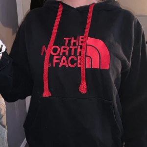 Svart hoodie från The North Face M - Svart hoodie från The North Face med rött tryck och röda snören. Klassisk känguruficka framtill och huva. Perfekt för chill dagar eller när du vill ha en streetstyle-look. Mjuk och skön, passar både tjejer och killar.