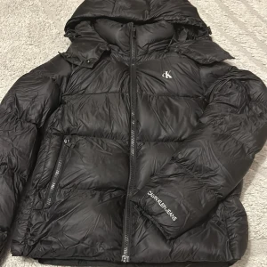 Svart pufferjacka från Calvin Klein Jeans - Svart pufferjacka från Calvin Klein Jeans med huva och dragkedja framtill. Jackan har logga på bröstet och ärmen samt stor logotyp på insidan. Perfekt för kalla dagar med sin vadderade design och snygga, stilrena look.