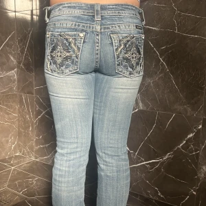 Miss Me ljusblå bootcut jeans 27 - Snygga ljusblå jeans från Miss Me med bootcut passform. Jeansen har slitningar framtill och coola broderade bakfickor med nitar och mönster. Klassisk femficksmodell med låg midja och detaljerade sömmar. Perfekta för dig som gillar statement-jeans.