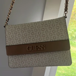 Beige axelväska från Guess - Snygg beige axelväska från Guess med kedjerem och logomönster över hela väskan. Väskan har ett stort Guess-emblem i guld på framsidan och stängs med magnetlås. Perfekt storlek för det viktigaste och ger en clean look.