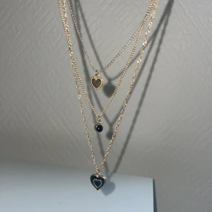 Guldigt lager-på-lager halsband med hjärtan - Trendigt lager-på-lager halsband i guld med tre kedjor. Ena hänget är ett litet guldhjärta, det andra en svart sten och det tredje ett större svart hjärta med guldkant. Perfekt accessoar för att lyfta din outfit.