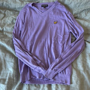 Lila v-ringad tröja från Lyle & Scott - Lila v-ringad tröja från Lyle & Scott med deras klassiska gula fågel-logga broderad på bröstet. Tröjan är långärmad och har ribbade muddar vid ärmslut och nederkant. Perfekt för dig som gillar stilrena och färgglada plagg.