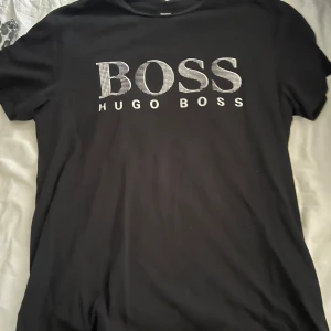 Svart t-shirt från Hugo Boss - Svart t-shirt från Hugo Boss med stort vitt BOSS-tryck på bröstet. Klassisk rund hals och korta ärmar. Skön och stilren modell som passar perfekt till jeans eller shorts. Materialet känns mjukt och följsamt. Är L men passar bra på en M.