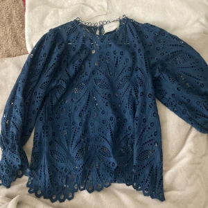 Blå spetsblus från Neo Noir - Supersnygg blå blus från Neo Noir med unikt broderat spetsmönster och vågig kant nertill. Blusen har långa ärmar och rund halsringning, perfekt för dig som gillar detaljer och vill sticka ut lite extra.