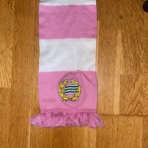 Rosa och vit Hammarby halsduk - Säljer en lång, stickad halsduk i rosa och vitt med breda ränder och Hammarby IF:s broderade klubbmärke. Halsduken har fransar i ändarna och är perfekt för att visa ditt stöd på matchen eller i stan. Lite sliten men fortfarande bra skick.
