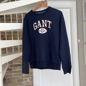 Mörkblå sweatshirt från GANT - Snygg mörkblå sweatshirt från GANT med broderad logga och text framtill. Klassisk rund halsringning och långa ärmar. Tröjan är i mjuk bomull och har ribbade muddar vid ärmslut och nederkant. Perfekt för en chill och sportig look.