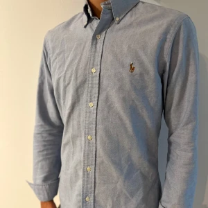 Blå skjorta från Polo Ralph Lauren - Klassisk ljusblå skjorta från Polo Ralph Lauren i slim fit-modell. Skjortan är tillverkad i 100% bomull, har knappar framtill och den ikoniska broderade loggan på bröstet. Perfekt för dig som gillar stilrena och tidlösa plagg.
