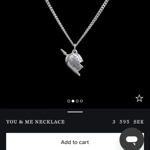 Maria Nilsdotter halsband  - Unikt halsband i silverfärg med ett coolt hjärtformat hänge som är delat och har en liten pil genom sig. Kedjan är grov och ger en trendig vibe. Perfekt att bära ensam eller att matcha med andra smycken för en personlig stil. 