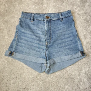 Blåa jeansshorts med uppvikt kant - Snygga blå jeansshorts med hög midja och två bakfickor. Shortsen har uppvikta benslut, bälteshällor och stängs med knapp och dragkedja. Perfekta för varma dagar och enkel att matcha med allt.