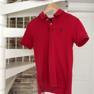 Röd pikétröja från Polo Ralph Lauren - Klassisk röd pikétröja från Polo Ralph Lauren med korta ärmar och krage. Tröjan har två knappar vid halsen och den ikoniska blå broderade loggan på bröstet. Tillverkad i mjuk bomull som är skön att bära hela dagen. De går att pruta 