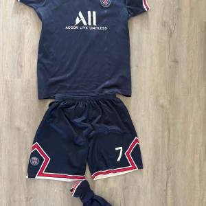 PSG Mbappé matchställ blå - Säljer ett komplett PSG matchställ med Mbappé 7-tryck. Tröjan är marinblå med vita och röda detaljer, klubbmärke på bröstet och sponsorloggor. Korta ärmar med randiga muddar, tillhörande shorts och strumpor. Tillverkat i lätt och ventilerande polyester.