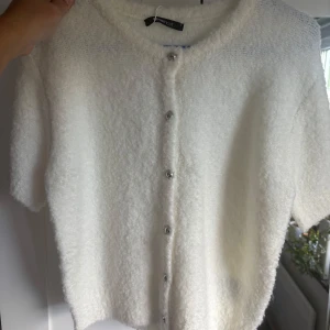 Vit fluffig kofta från Gina Tricot - Supermjuk och fluffig vit kofta från Gina Tricot med kort ärm och silvriga knappar framtill. Helt ny med prislapp kvar. 