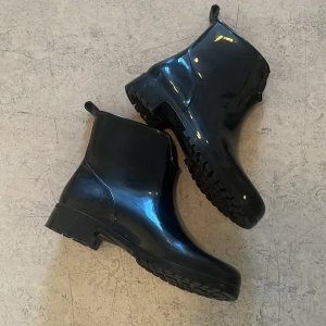 Svarta regn stövlar - Svarta boots med glansig finish och grov sula. Skorna är tillverkade i syntetmaterial och har en enkel, stilren design med rund tå. Perfekta för regniga dagar och ger en cool vibe till din outfit.