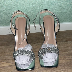  glittriga klackskor  - Säljer ett par Snygga klackskor i metallic silver med fyrkantig tå och hög klack. Remmarna runt vristen och över foten är dekorerade med glittrande stenar för extra bling. Aldrig använda, bara testade