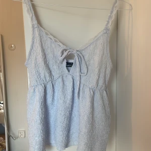 Ljusblå spetsdetaljerad linnetopp - Supersöt ljusblå linnetopp från Gina Tricot med spetskant längs axelbanden och v-ringning med knyt fram. Toppen har en krinklad struktur och är ärmlös, perfekt för varma dagar. Sitter löst och har en romantisk vibe.