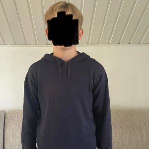 Mörkblå hoodie med huva - En basic mörkblå hoodie med huva och dragsko. Tröjan har lång ärm och en klassisk passform, perfekt för chill dagar. Materialet känns mjukt och skönt mot huden. Enkel och stilren design utan tryck eller logga.