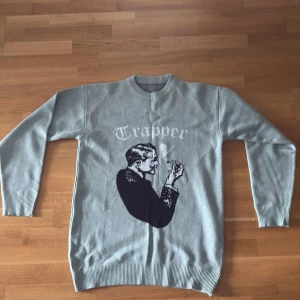 Ljusgrå stickad tröja med tryck Trapper - Ljusgrå stickad tröja med rund hals och ribbade muddar. På framsidan finns ett stort svartvitt motiv av en man som röker och texten 'Trapper' i vitt. Tröjan har lång ärm och en avslappnad passform, perfekt för dig som gillar unika prints.