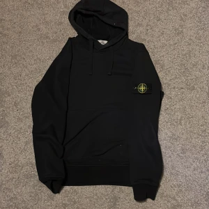 Svart hoodie från Stone Island - Svart hoodie från Stone Island med klassisk logga på ärmen. Tröjan har huva med snörning och en stor magficka framtill. Perfekt för dig som gillar streetwear och vill ha en stilren look. Materialet känns mjukt och skönt, troligen bomull.