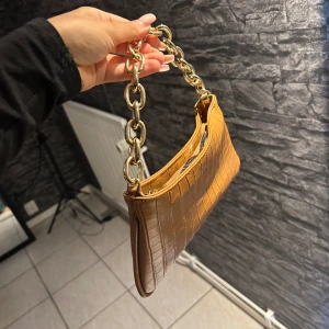 Brun axelväska med kedja - Snygg brun axelväska i croco-mönstrat skinnimitation med chunky guldfärgad kedja som handtag. Väskan har dragkedja upptill och ett stilrent innerfoder. Perfekt storlek för det viktigaste och ger en trendig vibe till din outfit.
