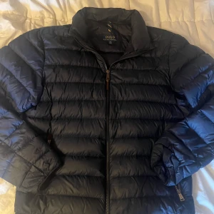 Mörkblå dunjacka Polo Ralph Lauren - Snygg mörkblå dunjacka från Polo Ralph Lauren i storlek M. Jackan har horisontella sömmar, hög krage och dragkedja framtill. Broderad logga på bröstet och två sidofickor med dragkedja. Perfekt för kyliga dagar.