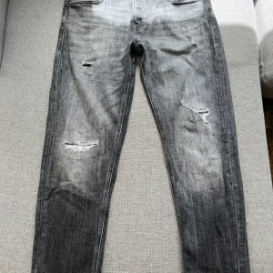 Grå slitna skinny jeans från Jack & Jones - Säljer ett par grå skinny jeans från Jack & Jones med slitningar och hål på benen för en riktigt edgy look. Jeansen har klassiska fem fickor, snygg tvätt och Jack & Jones signatur på bakfickan. Perfekta för dig som gillar streetstyle och vill sticka ut.