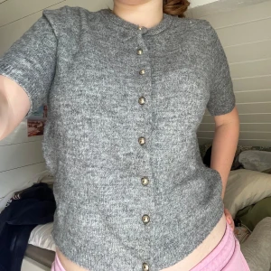 Grå stickad kofta från H&M - Kortärmad grå stickad kofta från H&M med rund hals och snygga guldfärgade knappar framtill. Koftan har ribbade muddar vid hals, ärmslut och nederkant. Perfekt att slänga över en topp för en chill och stilren look. Pris går stt diskuteras. 