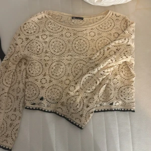 Beige spetsblus med virkat mönster - Superfin beige blus från Zara med virkat spetsmönster i cirklar och blommor. Croppad modell med långa ärmar och svarta detaljer längst ner. Perfekt för dig som gillar unika detaljer och boho-vibe.