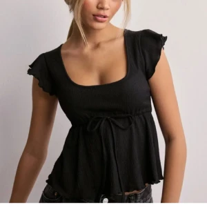 Svart blus med knyt från Nelly - Supersöt svart blus från Nelly med volangärmar, fyrkantig ringning och knyt i midjan. Toppen har lätt rynkad struktur och faller snyggt med en lite utsvängd nederdel. Perfekt att styla med jeans eller kjol för en trendig look.