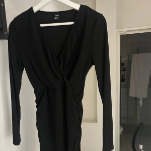 Svart omlottklänning från Shein - Snygg svart omlottklänning från Shein med v-ringning och långa ärmar. Klänningen har en figurnära passform och lätt rynkade detaljer i midjan. Perfekt för dig som gillar stilrena och trendiga plagg. 