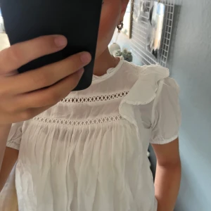 Vit blus med volang och spetsdetaljer - Supersöt vit blus med korta puffärmar, volang på axeln och broderade spetsdetaljer vid bröstet. Blusen är luftig och har en lös passform, perfekt till jeans eller kjol. Materialet känns lätt och somrigt, och den har en romantisk vibe.