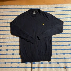 Svart stickad tröja från Lyle & Scott - Svart stickad tröja från Lyle & Scott med klassisk rund hals och gul broderad logga på bröstet. Tröjan har långa ärmar och ribbade muddar för en clean look. Perfekt för dig som gillar stilrena och tidlösa plagg.