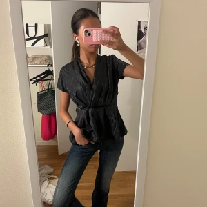 Svart omlottblus med kort ärm - Säljer en svart omlottblus med korta ärmar och markerad midja. Blusen har en lätt glansig yta och ett subtilt mönster i tyget. Den har v-ringning och voluminös peplum nedtill, vilket ger en snygg siluett. Perfekt att styla med jeans eller kjol.