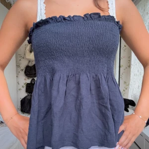 Mörkblå bandeau tubtopp med volang - Supersöt mörkblå bandeau tubtopp med smockad överdel och volangdetalj upptill. Toppen är ärmlös och har en luftig, lätt utställd passform nedtill. Perfekt för varma dagar och enkel att styla med jeans eller kjol.