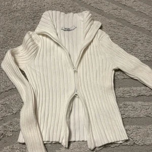 Vit ribbad kofta - Vit ribbad kofta med hög krage och hel dragkedja framtill. Tröjan är långärmad och har en tight passform som framhäver siluetten. Perfekt att styla med jeans eller kjol för en clean look.