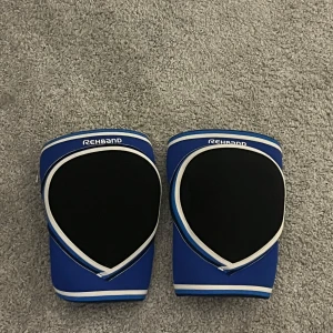 Blå träningsskydd från Rehband strl L - Säljer ett par blå träningsskydd från Rehband i storlek L. De har svart vaddering framtill och vita samt blå detaljer längs kanterna. Perfekta för dig som vill ha extra stöd vid träning eller sport. Snygg design och riktigt bekväma att använda.