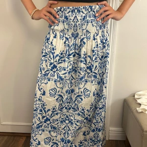 Vit och blå blommig långkjol/ klänning - Supersöt klänning som man kan ha som långkjol i vitt med blått blommönster och smockad midja. Kjolen är luftig och flowy, perfekt för varma dagar. Sitter bekvämt i midjan och har en riktigt somrig vibe. Passar dig som gillar boho-stil och vill sticka ut lite extra.