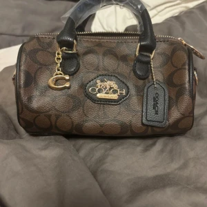 Brun handväska från Coach med monogram - Snygg brun handväska med klassiskt monogrammönster och svarta detaljer. Väskan har guldfärgade metalldetaljer, dragkedja och avtagbar axelrem. Perfekt storlek för det viktigaste och har Coach-logga framtill samt dekorativ C-berlock.