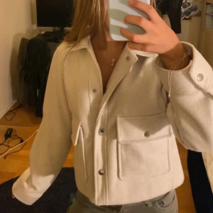 Beige kappa från Zara - Snygg beige kappa från Zara med stora fickor framtill och silvriga knappar. Jackan har klassisk krage och raka ärmar, perfekt att slänga över en hoodie eller t-shirt. Materialet känns mjukt och lite tjockare, vilket gör den extra mysig.