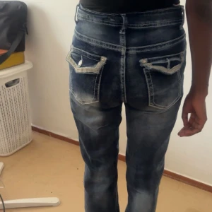Mörkblå högmidjade jeans med fickdetaljer - Snygga mörkblå jeans med hög midja och raka ben. Jeansen har slitningar och markerade sömmar samt unika bakfickor med lock och knapp. Materialet är klassisk denim och passformen är normal. Perfekta för dig som gillar en cool och avslappnad stil.
