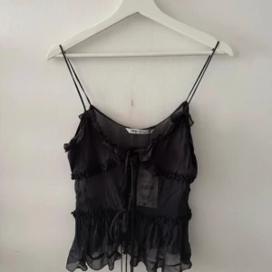 Svart volang topp från Zara  - Supersöt svart linnetopp från Zara med tunna axelband, volangdetaljer och knyt framtill. Toppen är endast testad och kappen är kvar.