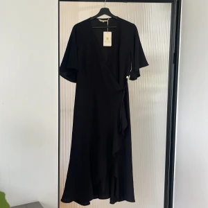Svart omlottklänning från By Pernilla Wahlgren - Supersnygg svart omlottklänning från med v-ringning, volangkant och korta vida ärmar. Klänningen har en dekorativ guldig knapp med ananasdetalj i sidan och knytband i midjan. Perfekt för dig som gillar stilrena och trendiga plagg. Ej byte/öppet köp. 