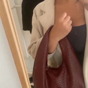 Vinröd axelväska i flätat skinn - Vinröd axelväska med snygg flätad struktur och rundad form. Väskan är tillverkad i skinnimitation och har dragkedja upptill. Perfekt storlek för att få plats med allt du behöver och ger en cool vibe till din outfit.