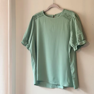 Grön blus i polyester - Säljer en stilren grön blus i 100% polyester. Blusen har korta ärmar med resår och en rund halsringning. 