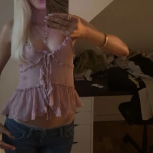 Rosa volangtop med knyt och tunna band - Supersöt ljusrosa linnetopp med volanger, tunna axelband och knyt framtill. Toppen har v-ringning, volangdetaljer längs bröstet och nederkanten samt en luftig, flowy passform. Perfekt för varma dagar och ger en romantisk vibe. Från Zara, slutsåld som ny 