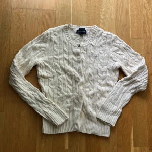 Vit kabelstickad kofta från Ralph Lauren - Kabelstickad vit kofta från Ralph Lauren med rund halsringning och knappar framtill. Tröjan har långa ärmar, ribbade muddar och diskret broderad logga på bröstet. Perfekt för lager-på-lager och klassisk preppy stil.