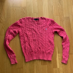 Rosa kabelstickad tröja från Ralph Lauren - Säljer en rosa kabelstickad tröja från Ralph Lauren med rund halsringning och diskret blå logga på bröstet. Tröjan har lång ärm och ribbade muddar, perfekt för dig som gillar klassisk preppy stil.